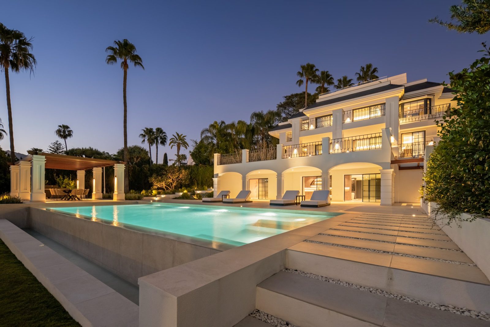 Illuminated villa with balconies, swimming pool, loungers and pergola at dusk in Marbella, Málaga - The Estates