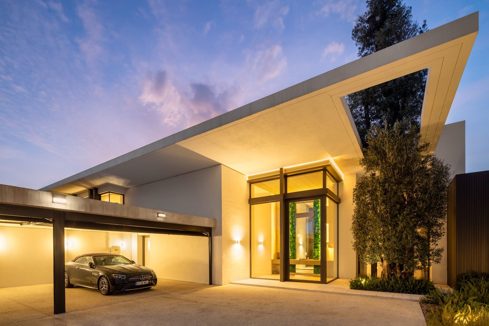 Modern villa entrance with illuminated carport, glass doorway and landscaped olive trees at dusk, of a 6-bedroom villa in Málaga - The Estates