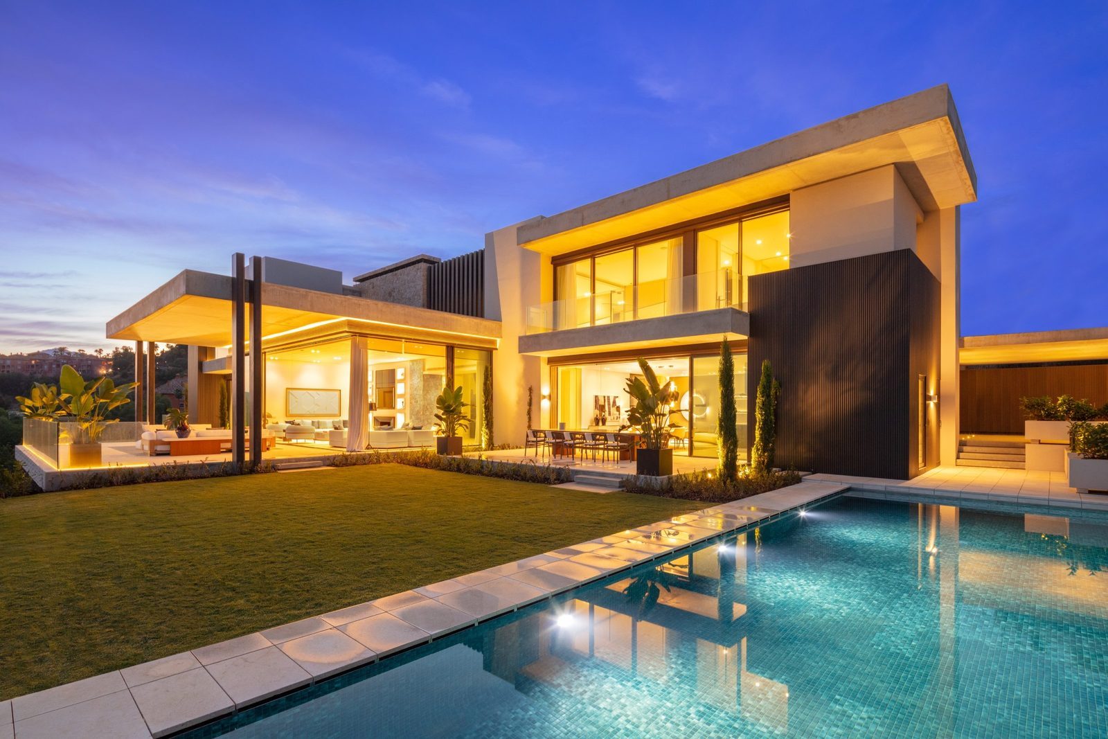 Modern villa exterior with illuminated interiors, landscaped garden and swimming pool at dusk in Málaga - The Estates