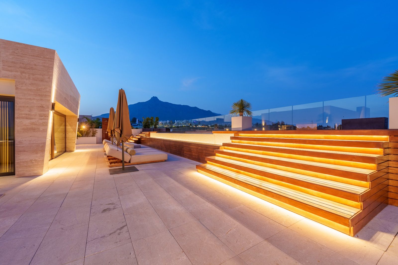 Illuminated rooftop terrace with loungers, umbrellas and stepped seating overlooking mountains of a 4