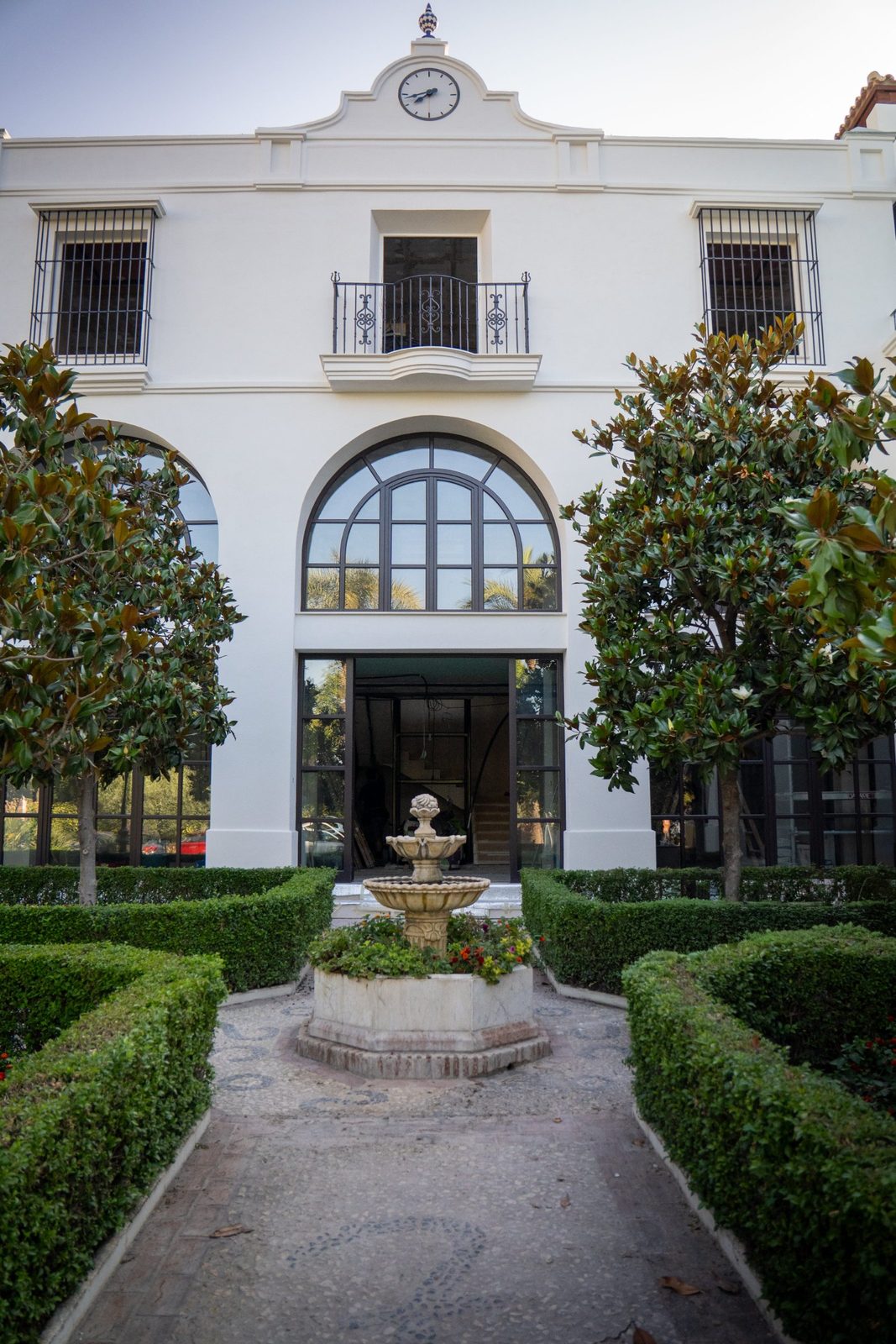 White villa facade with clock, arched windows, fountain and formal hedges in Benahavís, Málaga - The Estates