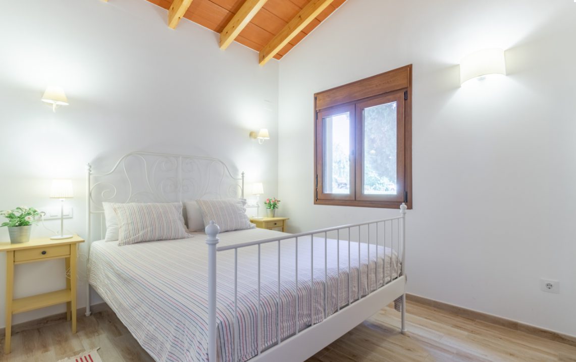 A bright bedroom with a wooden beam ceiling and window overlooking greenery in an 8-bedroom finca in Moraira - The Estates