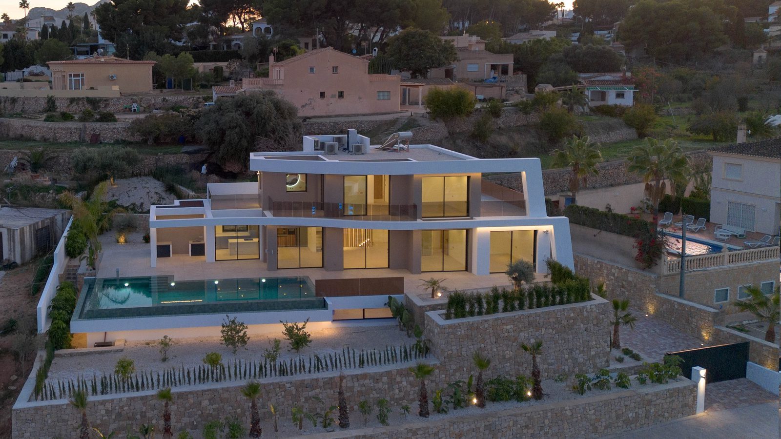 Modern four-bedroom villa with illuminated pool, terraces and landscaped gardens at dusk in Benissa - The Estates