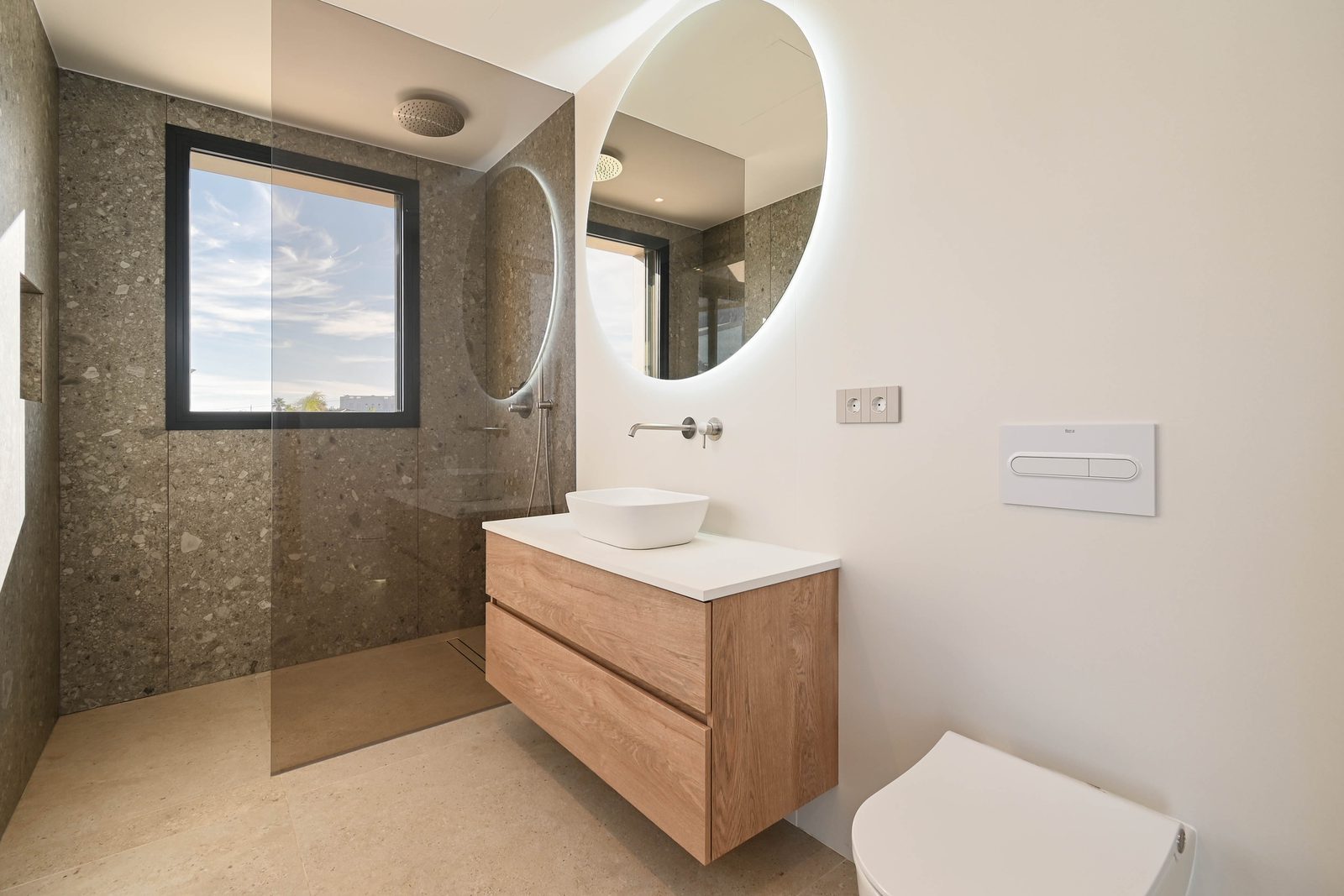 Modern bathroom with walk-in shower, stone-effect walls, floating wood vanity and round mirror in a 4-bedroom villa in Benissa - The Estates
