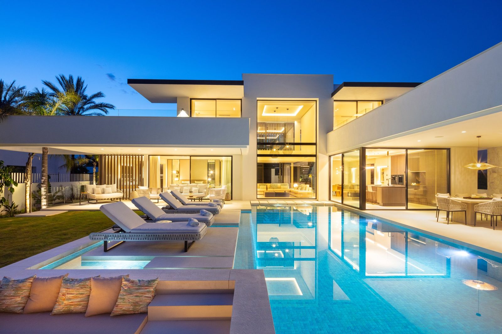 Modern villa exterior with illuminated interiors, pool, sun loungers and outdoor seating at dusk in Las Brisas, Marbella - The Estates
